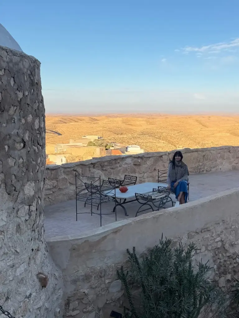 Bejal sitting on the terrace wearing a brown jumper and denim skirt with the sunrise behind her. There is a view of the desert highlands behind her
