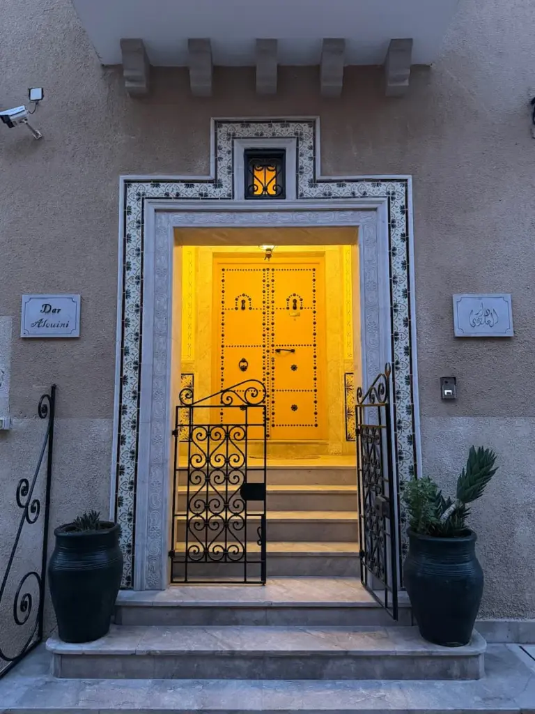 Dar Alouini front door in Kairouan, Tunisia. The door is yellow and studded in black. There are plant pots at either side of the door which has a light on in the background