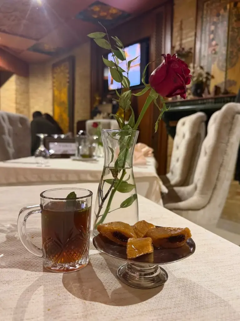 A plate of Makroudh at El Brija, Kairouan, Tunisia next to a glass of mint tea on a table with a white table cloth and other diners in the background. There is also a red rose in a small vase next to the Makroudh